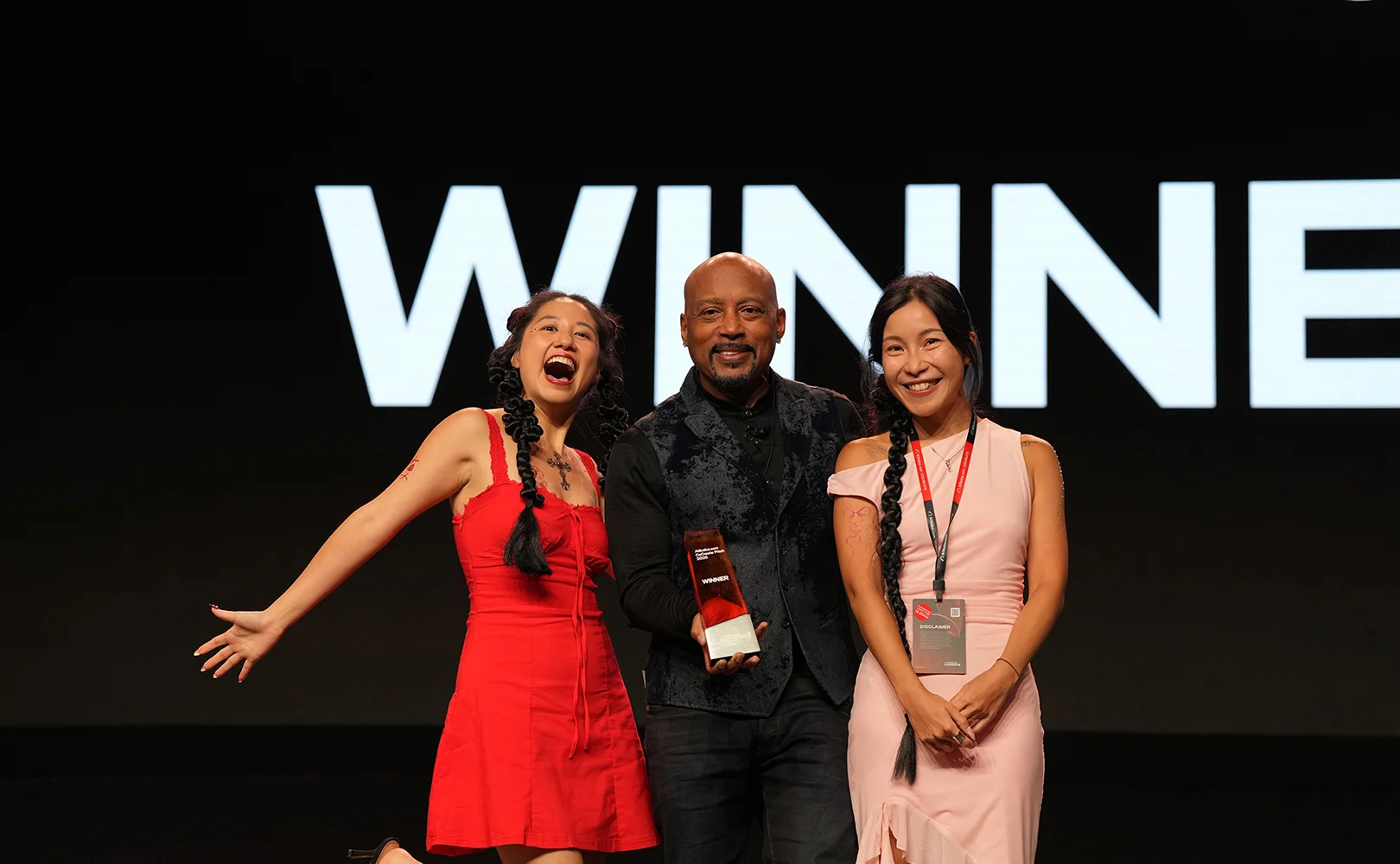 Two CoCreate Pitch 2025 winners on stage holding their award trophy alongside host Daymond John, with the word WINNER projected on the backdrop behind them.
